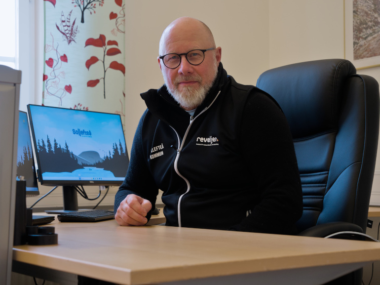 Man med glasögon sitter vid skrivbord. I bakgrunden syns datorskärmar. 