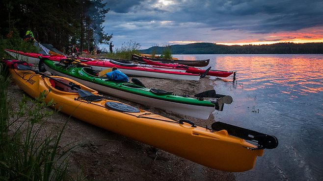 Bilden visar flera kanoter uppradade på en strand i Ångermanälven, solen håller på att gå ner